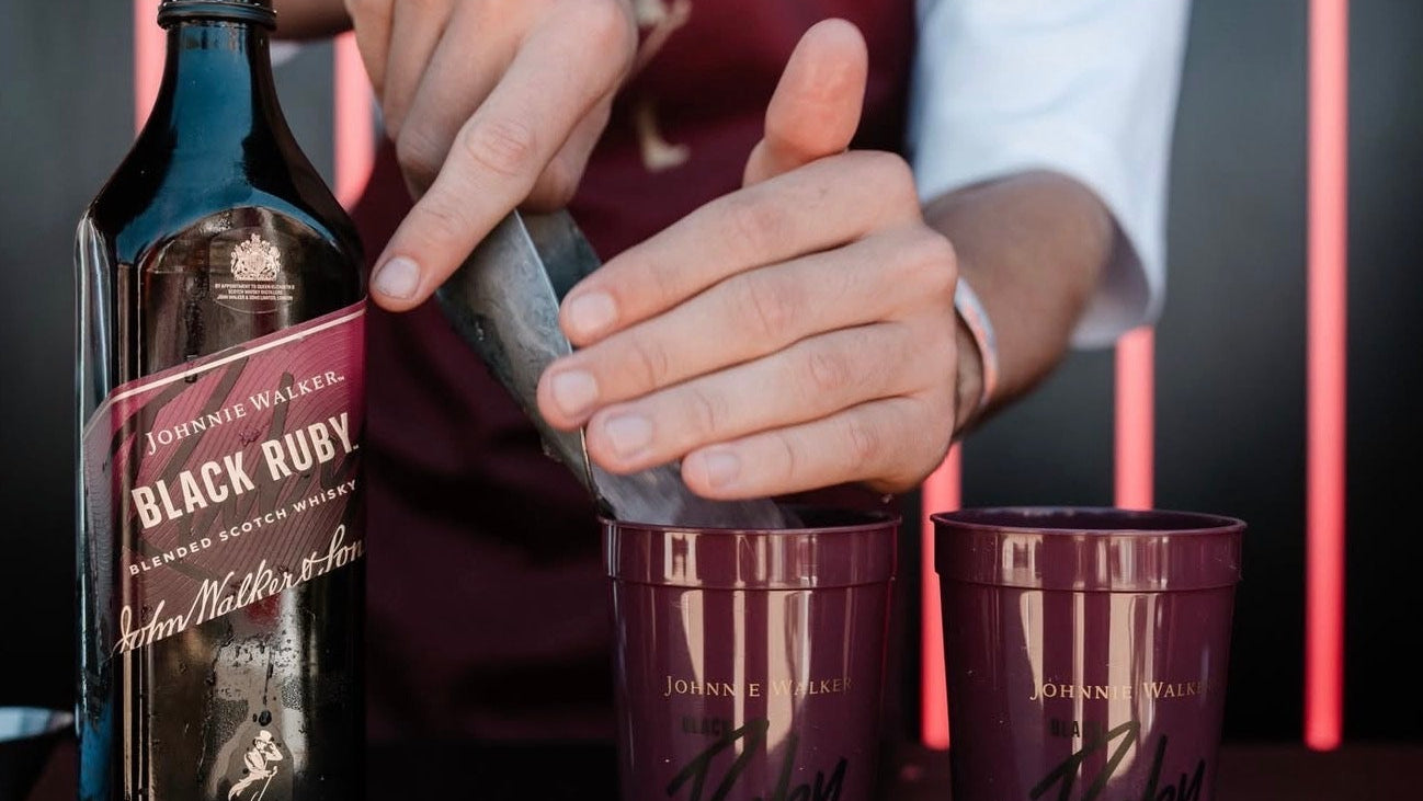 Person in a maroon apron preparing a drink with Johnnie Walker Black Ruby bottle and cups.
